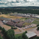 Vue aérienne d'un grand complexe immobilier avec plusieurs parkings, entouré d'arbres, de champs et de collines lointaines, sous un ciel partiellement nuageux. Plusieurs voitures sont garées et une voie ferrée longe le bord inférieur.