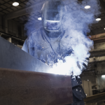 Une personne portant un équipement de protection, notamment un casque de soudage, soude une grande poutre métallique dans un environnement industriel. Des étincelles brillantes et de la fumée sont visibles autour de la zone de soudage.