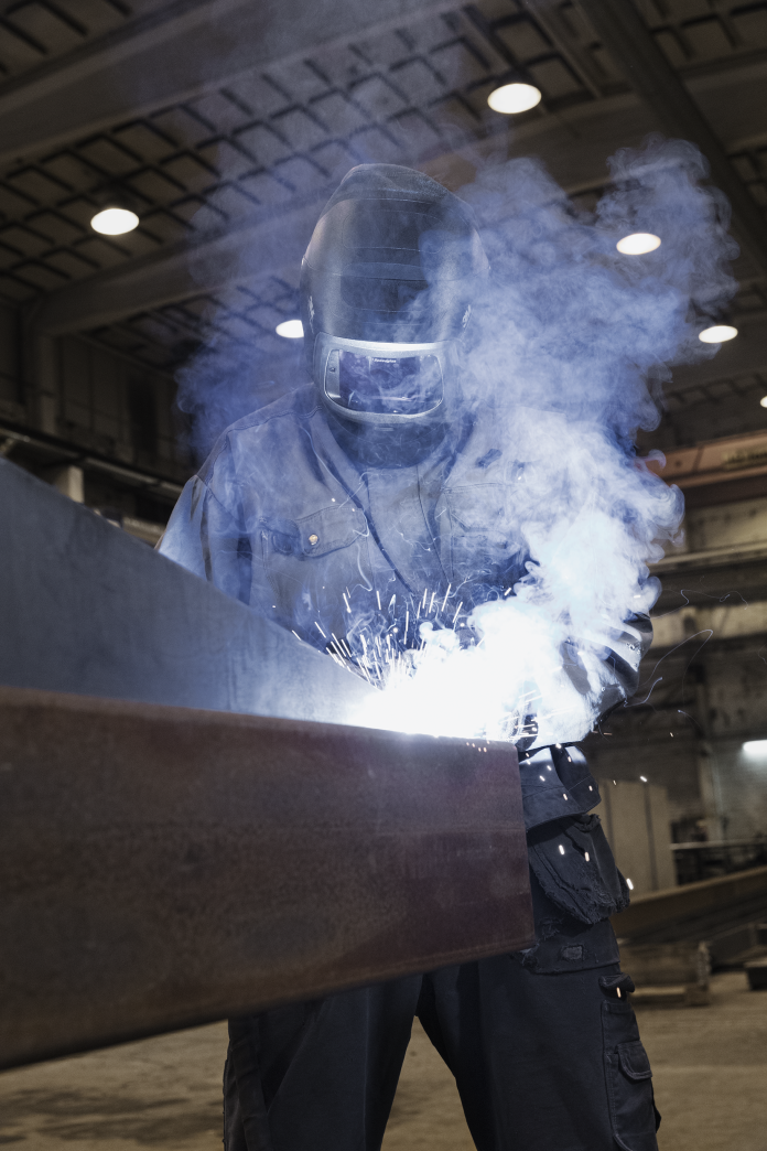 Une personne portant un équipement de protection, notamment un casque de soudage, soude une grande poutre métallique dans un environnement industriel. Des étincelles brillantes et de la fumée sont visibles autour de la zone de soudage.