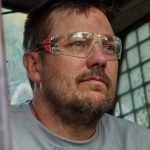 Un homme aux cheveux courts, barbu et portant des lunettes de sécurité est assis à l'intérieur d'un véhicule à cage métallique, peut-être un engin de chantier, et regarde à l'extérieur avec une expression concentrée.