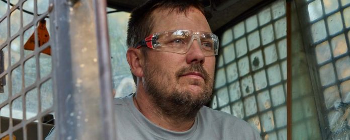 Un homme aux cheveux courts, barbu et portant des lunettes de sécurité est assis à l'intérieur d'un véhicule à cage métallique, peut-être un engin de chantier, et regarde à l'extérieur avec une expression concentrée.