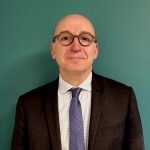 Un homme d'âge moyen avec des lunettes, portant un costume sombre, une chemise blanche et une cravate bleue à motifs, se tient devant un mur sarcelle uni, regardant la caméra avec une expression neutre.