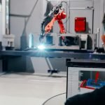 Une personne utilise un ordinateur portable avec un logiciel d'ingénierie affichant un modèle 3D tandis qu'un bras robotisé travaille sur une machine en arrière-plan, émettant une lumière vive dans un environnement industriel.