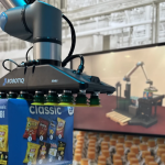 Un bras robotisé doté d'une pince aspirante tient une boîte de chips assorties ; un écran à l'arrière-plan affiche un graphique d'un bras robotisé similaire dans un entrepôt.