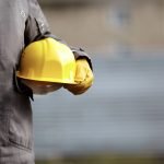 Une personne portant une veste de travail grise et des gants jaunes tient un casque de chantier jaune à ses côtés. L'arrière-plan est flou.