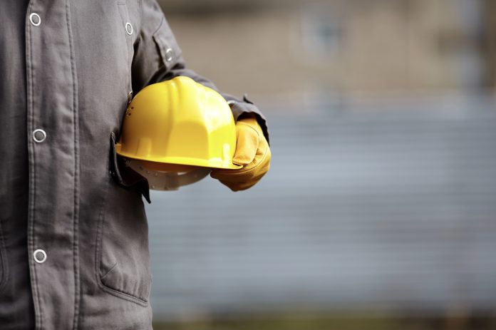 Une personne portant une veste de travail grise et des gants jaunes tient un casque de chantier jaune à ses côtés. L'arrière-plan est flou.