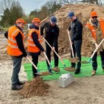 Cinq hommes, dont quatre portent des gilets de sécurité orange et des casques de protection, se tiennent sur un chantier de construction. Ils utilisent des pelles pour creuser dans un tas de terre devant eux, avec un tapis vert et un bloc de béton sur le sol.