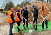 Modernisation de l’usine Trumpf à Haguenau Cinq hommes, dont quatre portent des gilets de sécurité orange et des casques de protection, se tiennent sur un chantier de construction. Ils utilisent des pelles pour creuser dans un tas de terre devant eux, avec un tapis vert et un bloc de béton sur le sol.