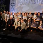 Un grand groupe de personnes, certaines debout et d'autres agenouillées, posent ensemble sur une estrade en tenant des prix. Une bannière en arrière-plan indique "SIMODEC". La plupart des personnes sont souriantes et plusieurs d'entre elles portent un badge de la conférence.