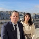 Un homme en costume et une femme en pull léger sont assis côte à côte sur une corniche en pierre, partageant un rendez-vous tranquille avec des immeubles parisiens et un ciel bleu en arrière-plan, évoquant l'esprit de Global Industrie.