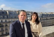 Un homme en costume et une femme en pull léger sont assis côte à côte sur une corniche en pierre, partageant un rendez-vous tranquille avec des immeubles parisiens et un ciel bleu en arrière-plan, évoquant l'esprit de Global Industrie.