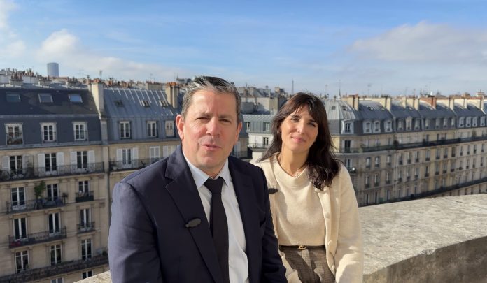 Un homme en costume et une femme en pull léger sont assis côte à côte sur une corniche en pierre, partageant un rendez-vous tranquille avec des immeubles parisiens et un ciel bleu en arrière-plan, évoquant l'esprit de Global Industrie.