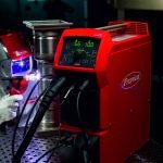 Un poste de soudage Fronius rouge avec écran numérique et câbles connectés est posé sur une table de travail métallique. À l'arrière-plan, une personne en tenue de protection soude à proximité d'un élément métallique.