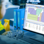 Dans un atelier ou un bureau, une personne est assise à un bureau et regarde un écran d'ordinateur affichant un logiciel de CAO ou d'ingénierie avec des diagrammes, des tableaux de données et des spécifications de pièces.