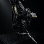 Un bras robotisé recouvert d'une gaine de protection noire, équipé d'une torche de soudage, se tient sur une surface sombre et nervurée sur fond noir.