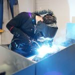 Une personne portant un équipement de protection, notamment un casque de soudage et des gants, soude des pièces métalliques sur un poste de travail, des étincelles vives et de la lumière étant visibles lors du processus de soudage.