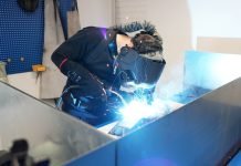 Une personne portant un équipement de protection, notamment un casque de soudage et des gants, soude des pièces métalliques sur un poste de travail, des étincelles vives et de la lumière étant visibles lors du processus de soudage.