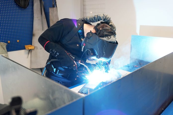 Une personne portant un équipement de protection, notamment un casque de soudage et des gants, soude des pièces métalliques sur un poste de travail, des étincelles vives et de la lumière étant visibles lors du processus de soudage.