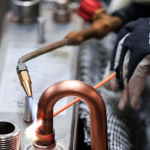 Une personne portant des gants de protection utilise un chalumeau pour souder ou braser des tuyaux en cuivre sur une structure métallique dans un environnement industriel. Divers outils et composants métalliques sont visibles à l'arrière-plan.