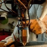 Une personne portant un équipement de protection utilise ses deux mains pour faire fonctionner une machine industrielle, en guidant une buse métallique dans un récipient rond dans un atelier ou une usine.