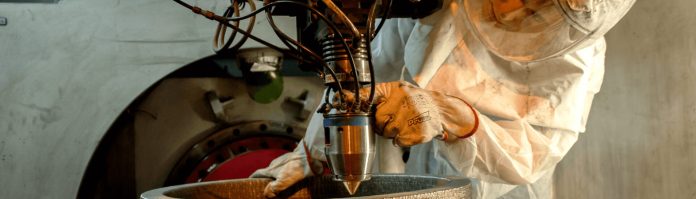 Une personne portant un équipement de protection utilise ses deux mains pour faire fonctionner une machine industrielle, en guidant une buse métallique dans un récipient rond dans un atelier ou une usine.