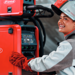 Une personne portant un équipement de sécurité et des gants rouges utilise une machine à souder Fronius et ajuste le panneau de commande tout en souriant et en regardant la caméra.