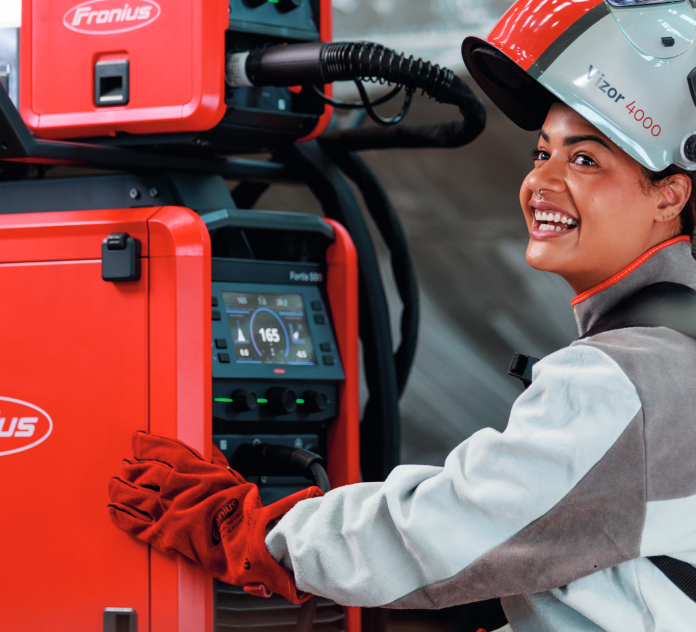Une personne portant un équipement de sécurité et des gants rouges utilise une machine à souder Fronius et ajuste le panneau de commande tout en souriant et en regardant la caméra.