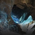 Une personne portant un casque et une veste de protection pour le soudage est en train de souder du métal, des étincelles et de la fumée étant visibles autour de ses mains. Le mot "bolle" est imprimé sur le devant du casque.