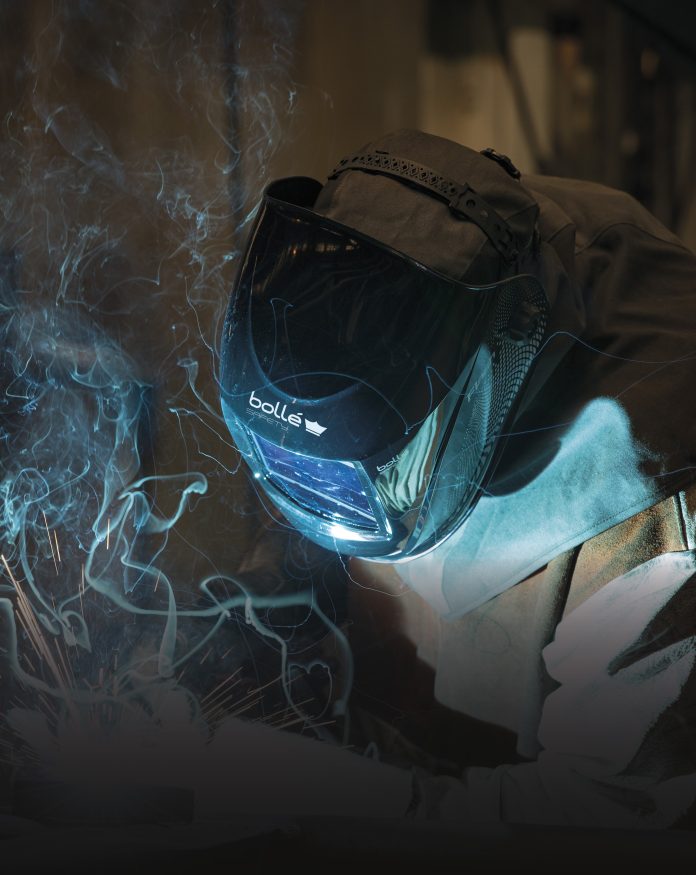 Une personne portant un casque et une veste de protection pour le soudage est en train de souder du métal, des étincelles et de la fumée étant visibles autour de ses mains. Le mot "bolle" est imprimé sur le devant du casque.