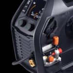 Vue rapprochée d'une machine à souder noire avec deux boutons verts, un écran numérique indiquant les réglages, des boutons de réglage, et divers connecteurs et commandes sur le panneau avant.