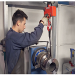 Une personne portant des gants de travail et un tablier sombre actionne un appareil de levage rouge pour déplacer une grande pièce métallique dans un atelier industriel. Des machines et des panneaux de sécurité sont visibles à l'arrière-plan.