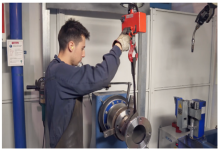 Une personne portant des gants de travail et un tablier sombre actionne un appareil de levage rouge pour déplacer une grande pièce métallique dans un atelier industriel. Des machines et des panneaux de sécurité sont visibles à l'arrière-plan.