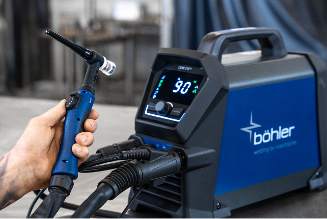 Une personne tient une torche de soudage bleue et noire connectée à un poste de soudage TIG Böhler générateurs CORE bleu affichant "90" sur son écran numérique. L'appareil se trouve sur un plan de travail à l'intérieur.