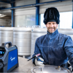 Un homme portant des équipements de soudage et un casque de soudure levé sourit alors qu'il est assis à un poste de travail avec une machine à souder CORE Bohler et des réservoirs métalliques en arrière-plan, reflétant un environnement professionnel.