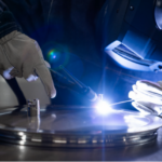 Une personne portant des gants et un casque de soudage utilise une torche TIG de soudage sur un objet métallique rond, avec une grande précision car un arc lumineux bleu brille au point de contact.