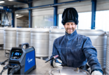 Un homme portant des équipements de soudage et un casque de soudure levé sourit alors qu'il est assis à un poste de travail avec une machine à souder CORE Bohler et des réservoirs métalliques en arrière-plan, reflétant un environnement professionnel.