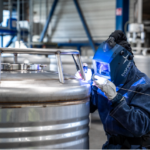 Une personne portant un équipement de protection et un casque de soudage effectue un soudage TIG avec une grande précision sur le joint d'une grande cuve industrielle en acier inoxydable dans une usine.