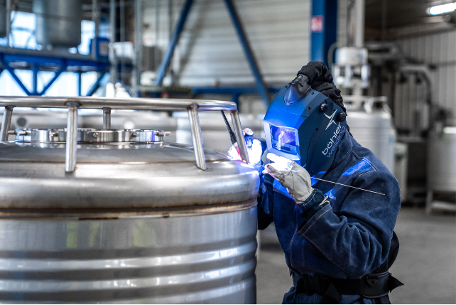 Une personne portant un équipement de protection et un casque de soudage effectue un soudage TIG avec une grande précision sur le joint d'une grande cuve industrielle en acier inoxydable dans une usine.