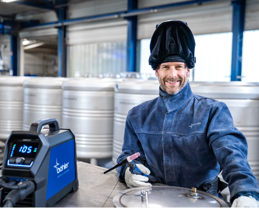 Un homme portant des équipements de soudage et un casque de soudure levé sourit alors qu'il est assis à un poste de travail avec une machine à souder CORE Bohler et des réservoirs métalliques en arrière-plan, reflétant un environnement professionnel.