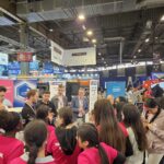 Un groupe de personnes, dont plusieurs filles portant des chemises roses, écoutent des présentateurs dans un hall d'exposition très fréquenté, rempli de stands industriels, d'enseignes et de machines.