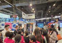 Global Industrie 2026 : mobilisation pour l’industrie française Un groupe de personnes, dont plusieurs filles portant des chemises roses, écoutent des présentateurs dans un hall d'exposition très fréquenté, rempli de stands industriels, d'enseignes et de machines.
