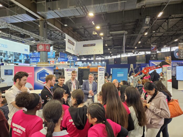 Un groupe de personnes, dont plusieurs filles portant des chemises roses, écoutent des présentateurs dans un hall d'exposition très fréquenté, rempli de stands industriels, d'enseignes et de machines.