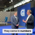 Deux hommes en tenue de travail se tiennent debout et discutent dans un hall d'exposition moderne à la décoration bleue et grise. Un grand logo "G" est affiché sur un mur derrière eux. La légende indique : "Ils sont venus en nombre.