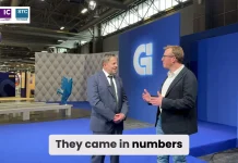 Deux hommes en tenue de travail se tiennent debout et discutent dans un hall d'exposition moderne à la décoration bleue et grise. Un grand logo "G" est affiché sur un mur derrière eux. La légende indique : "Ils sont venus en nombre.