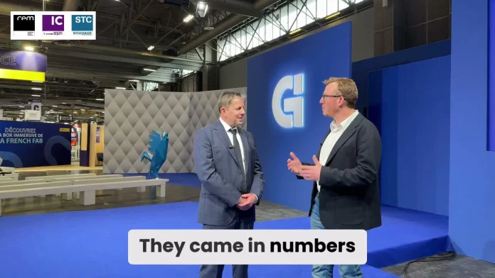 Deux hommes en tenue de travail se tiennent debout et discutent dans un hall d'exposition moderne à la décoration bleue et grise. Un grand logo 