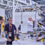 Deux hommes se tiennent près d'un bras robotique industriel dans une usine ou un laboratoire, observant son fonctionnement. D'autres personnes travaillent à l'arrière-plan. Des machines et des équipements sont visibles dans tout l'espace.