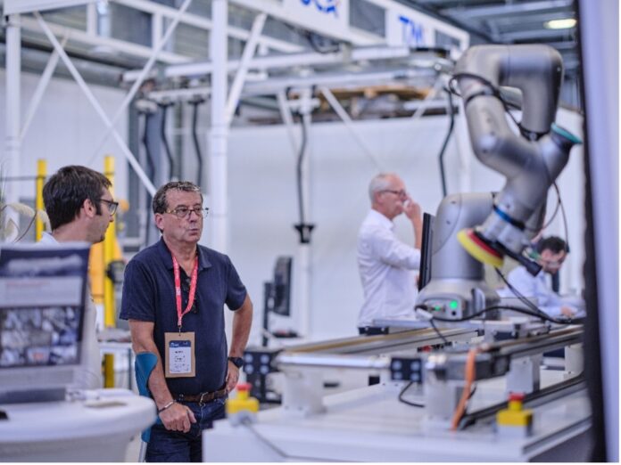 Deux hommes se tiennent près d'un bras robotique industriel dans une usine ou un laboratoire, observant son fonctionnement. D'autres personnes travaillent à l'arrière-plan. Des machines et des équipements sont visibles dans tout l'espace.