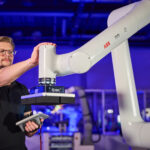 Une personne fait fonctionner un grand bras robotisé blanc étiqueté "ABB" dans un environnement industriel, à l'aide d'un dispositif de commande portable. Le bras robotique tient un objet plat et noir. L'arrière-plan montre des machines et un éclairage bleu.
