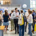 Un groupe de personnes se promène et discute parmi les stands d'un salon de la création d'entreprise. Des bannières et des présentoirs colorés entourent la foule, et certains participants portent des sacs fourre-tout et des badges nominatifs.
