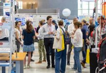 Un groupe de personnes se promène et discute parmi les stands d'un salon de la création d'entreprise. Des bannières et des présentoirs colorés entourent la foule, et certains participants portent des sacs fourre-tout et des badges nominatifs.